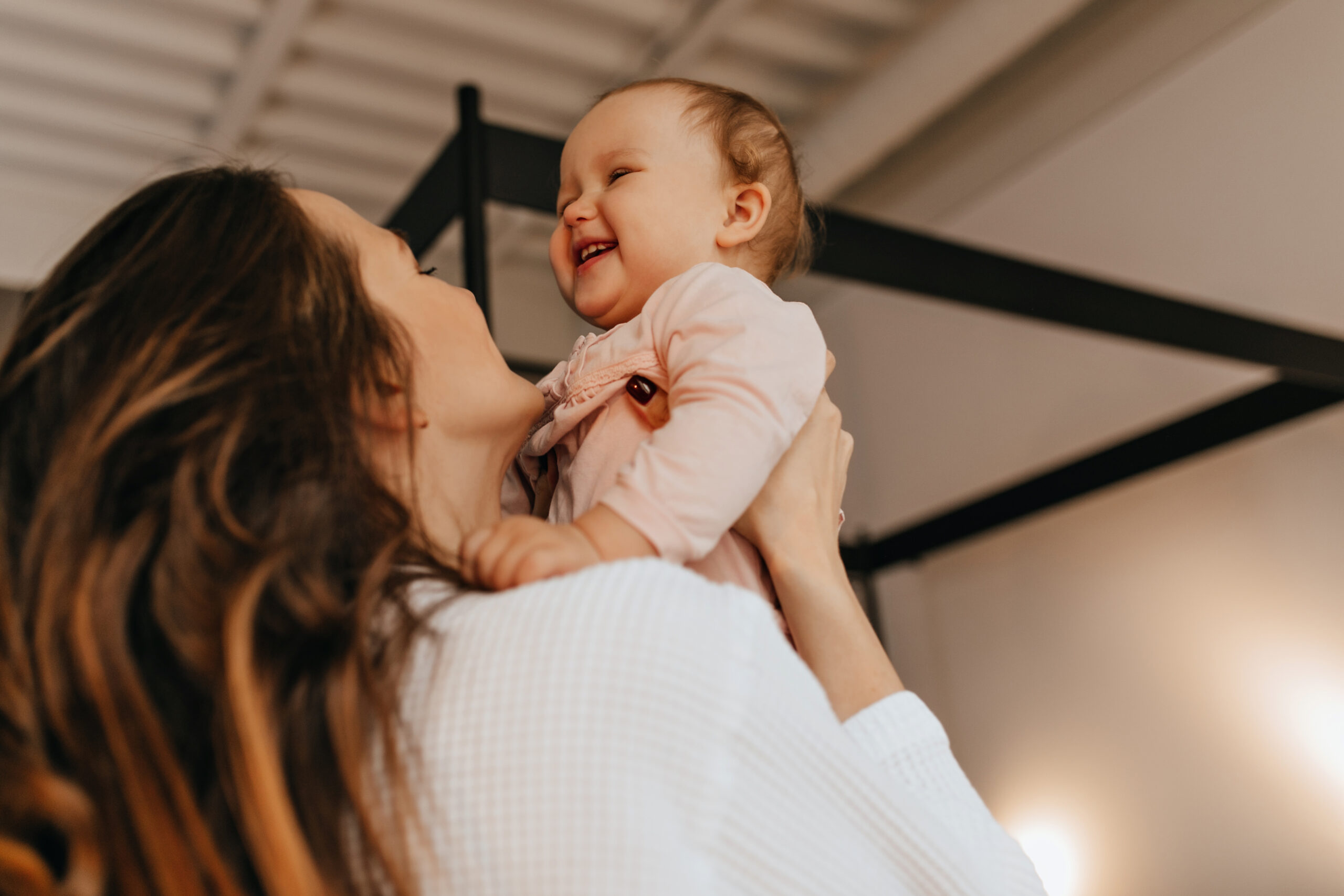 woman with long hair tickles and throws up laughing baby in light colored home sweater.